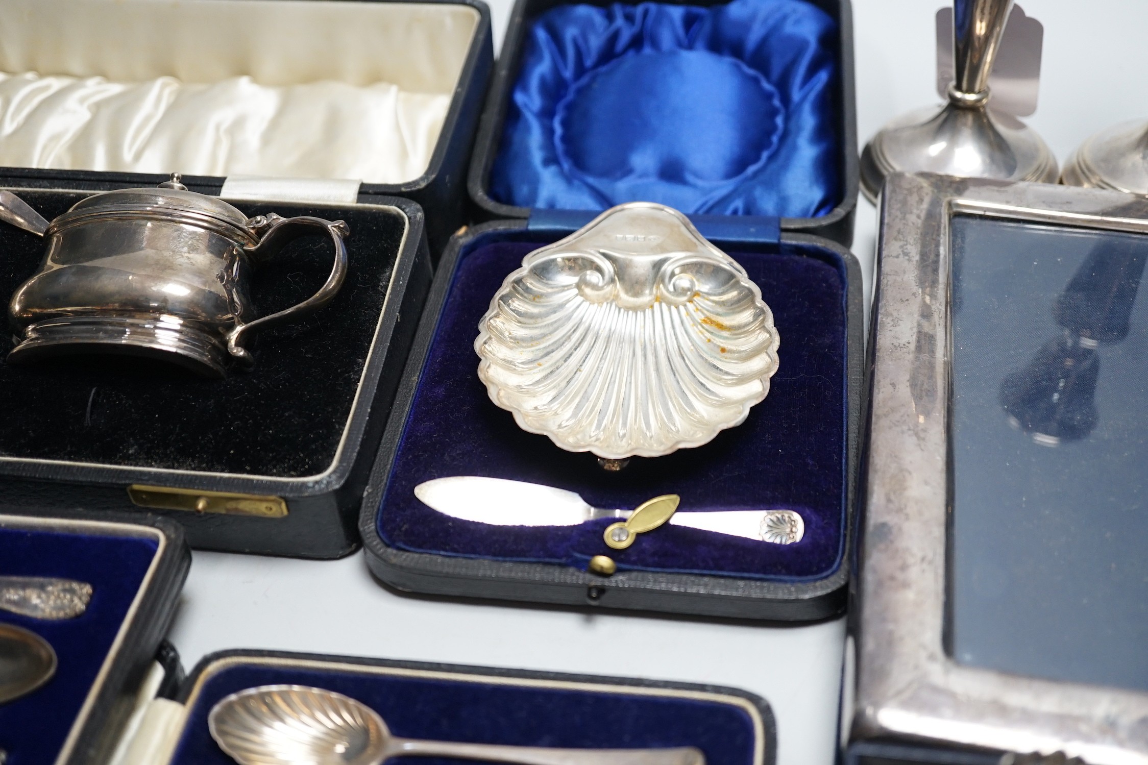 A quantity of mixed silver ware including a pair of modern candlesticks, 15cm, weighted, a cased George V three piece condiment set, cased butter shell and knife, two cased sets of teaspoons, a mounted photograph frame,